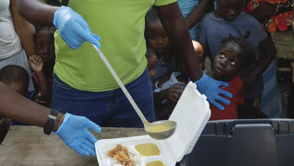 a server ladles soup into a container as children line up to receive food at a shelter for families displaced by gang violence in port au prince haiti march 14 2024