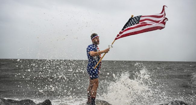 Youtuber mark peyton holds a us flag as he poses for his brother matt peyton on the shoreline ahead