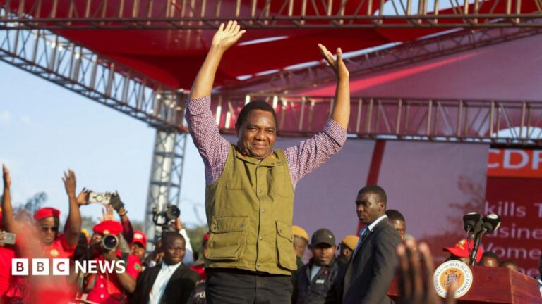 Zambia's President Hakainde Hichilema in green half-coat and black trousers lifts his hands to greet United Party for National Development supporters