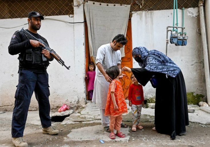PAKISTAN-HEALTH-POLIO-VACCINE This photograph taken on October 29, 2024 shows an elite police personnel (L) standing guard as a health worker (R) administers polio drops to a child during a door-to-door poliovirus vaccination campaign on the outskirts of Peshawar. - Pakistan has witnessed a dramatic uptick in attacks since the return of the Taliban in neighbouring Afghanistan in 2021, with Islamabad claiming hostile groups are now operating from there. Militant attacks and suspicion stemming from misinformation are hampering Pakistan's battle to eradicate polio, but teams of dedicated volunteer health workers are determined to fight on. (Photo by Abdul MAJEED / AFP) / TO GO WITH 'PAKISTAN-HEALTH-POLIO-VACCINE, FOCUS' BY LEHAZ ALI, WITH JULIETTE MANSOUR
